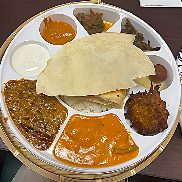 A Spicy Chowk Indian thali with various curries, rice, papadum, and dessert on a white segmented plate.