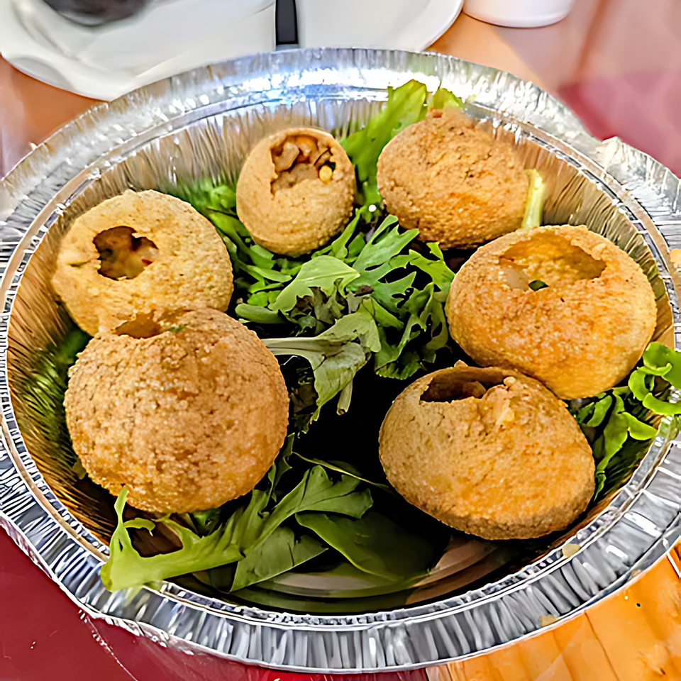Six Indian pani puri, some filled, on a bed of greens in a foil container.