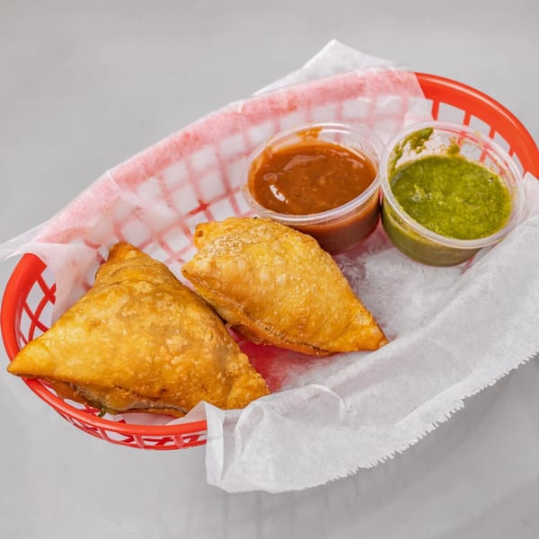 Two golden-brown samosas in a red basket with white paper, accompanied by two dipping sauces.